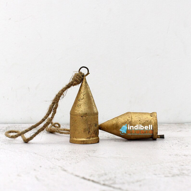 May include: Two antique brass-colored bells with a rustic rope hanging from one. The bells have a conical shape and a weathered finish, suggesting age and use. The bells are resting on a white surface.