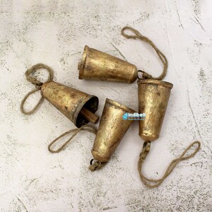 Puede incluir: Cuatro campanas de metal de color dorado con un cordel de yute atado. Las campanas est&aacute;n dispuestas en un racimo sobre una superficie blanca texturizada.