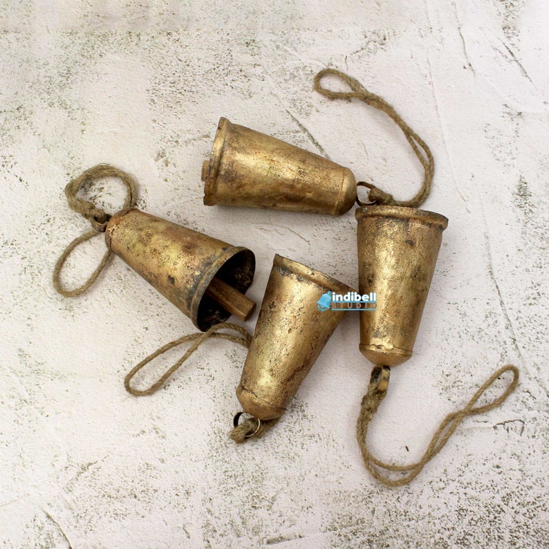 Puede incluir: Cuatro campanas de metal de color dorado con un cordel de yute atado. Las campanas est&aacute;n dispuestas en un racimo sobre una superficie blanca texturizada.