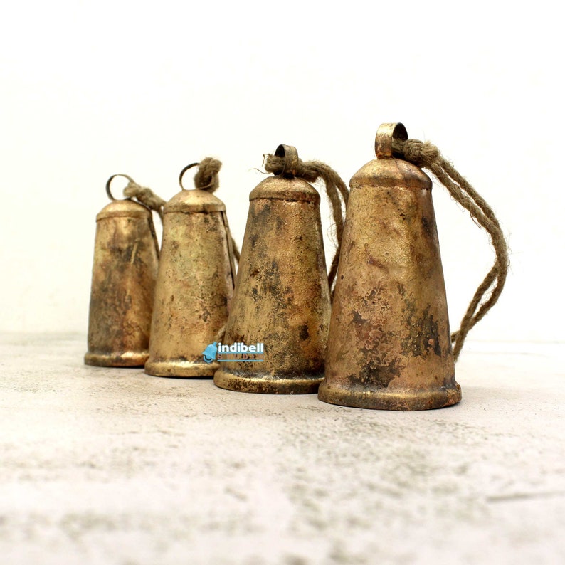 Puede incluir: Cuatro campanas de metal de color dorado con un acabado desgastado y una cuerda atada a cada una. Las campanas est&aacute;n dispuestas en fila sobre una superficie gris claro.