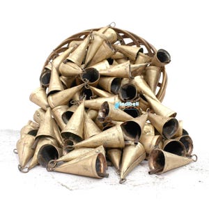 May include: A large collection of conical brass bells with a weathered gold finish, each featuring a small loop at the top and a clapper inside. The bells are piled together in a circular woven basket, with the text indibell studio visible.