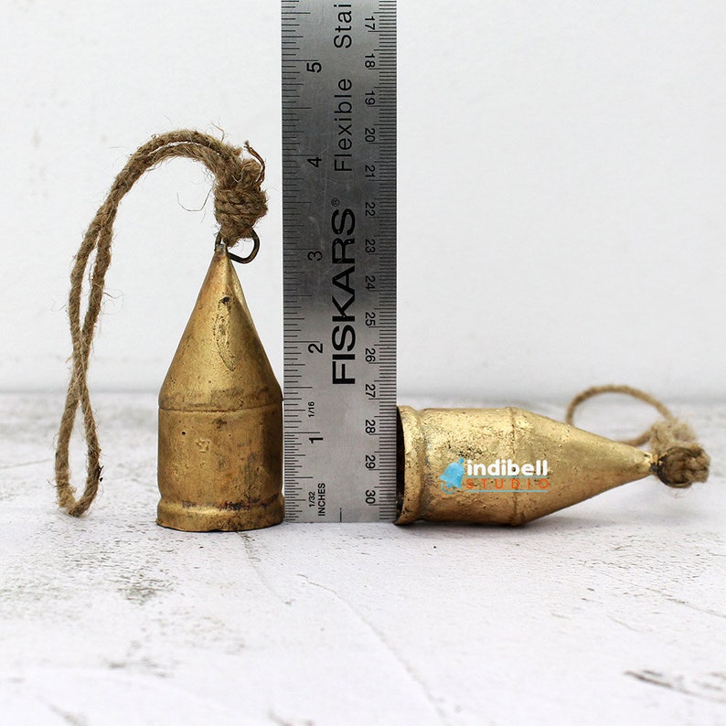 May include: Two brass-colored bells with a rope loop for hanging. One bell is upright, the other is lying down. The bells are approximately 3 inches tall. The bells have a rustic, vintage appearance.