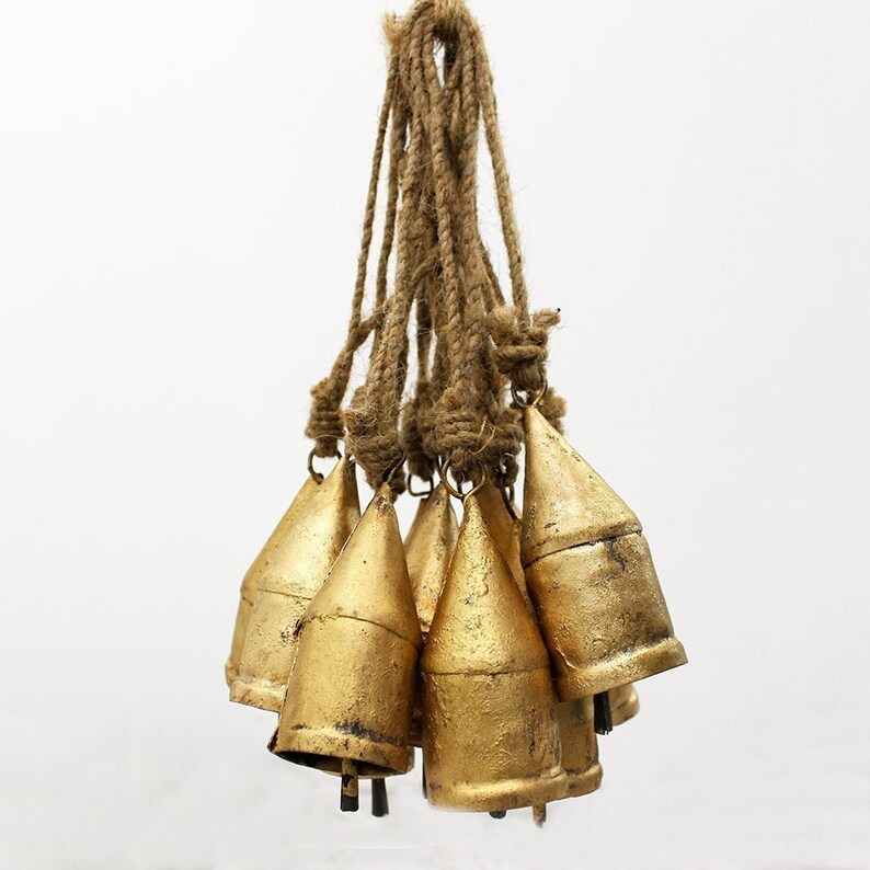 May include: A cluster of seven gold-colored metal bells hanging from a natural rope. The bells have a conical shape and a slightly distressed finish, suggesting a vintage or rustic aesthetic. The rope is thick and knotted.