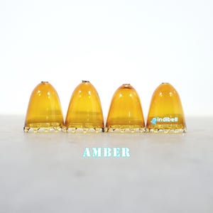 K&ouml;nnte beinhalten: Vier bernsteinfarbene, glockenf&ouml;rmige Glasabdeckungen mit gewelltem Rand. Jedes St&uuml;ck hat ein kleines Loch an der Oberseite. Der Text indibell ist auf der rechten Abdeckung aufgedruckt, das Wort AMBER steht in Hellblau unter den Objekten.