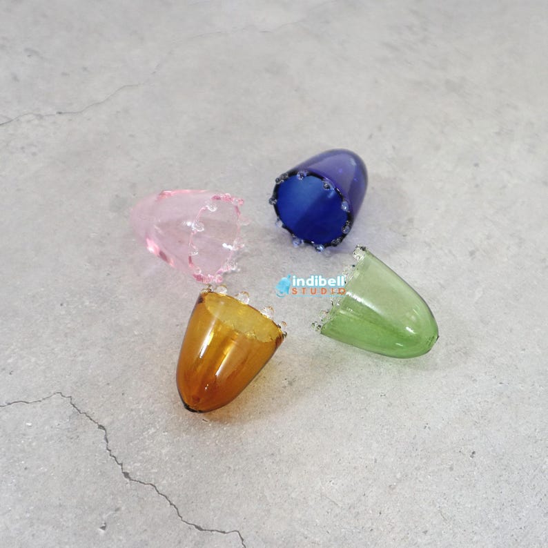 K&ouml;nnte beinhalten: Vier durchscheinende, kegelf&ouml;rmige Fingerh&uuml;te aus Glas in Rosa, Blau, Bernstein und Gr&uuml;n, jeweils mit einem dekorativen Perlenrand. Der Text indibell STUDIO ist in Blau und Orange in der Mitte der Anordnung aufgedruckt.