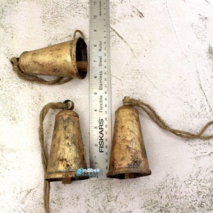 Puede incluir: Tres campanas de metal de color dorado con un acabado r&uacute;stico y cuerdas de cuerda. Las campanas miden aproximadamente 5 cm de alto y est&aacute;n dispuestas sobre una superficie blanca texturizada.