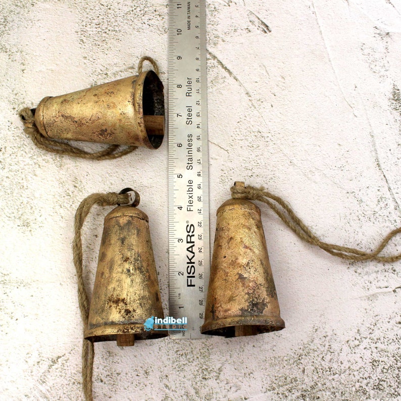 Puede incluir: Tres campanas de metal de color dorado con un acabado r&uacute;stico y cuerdas de cuerda. Las campanas miden aproximadamente 5 cm de alto y est&aacute;n dispuestas sobre una superficie blanca texturizada.