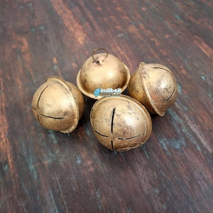 Può includere: Quattro campane antiche color oro con finitura anticata. Ogni campana ha un design a fessura a forma di croce e un piccolo anello nella parte superiore. Le campane sono disposte su una superficie scura con venature del legno.