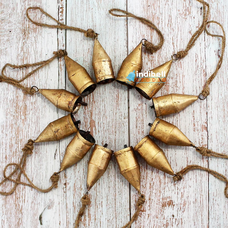 May include: A collection of ten golden-colored metal bells arranged in a circle. Each bell has a conical shape and is suspended by a natural-colored rope. The bells are displayed on a white wooden surface.