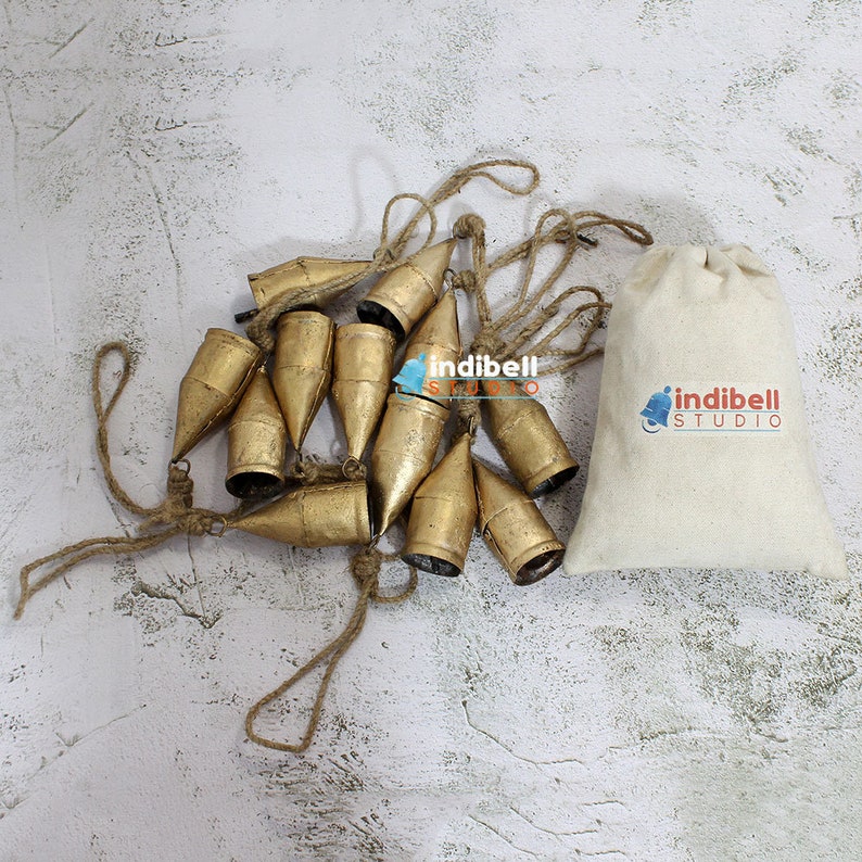 May include: A collection of gold-colored metal bells strung on a natural rope, with a matching drawstring bag. The bells have a conical shape and are arranged in a cluster. The bag has the "indibell STUDIO" logo.