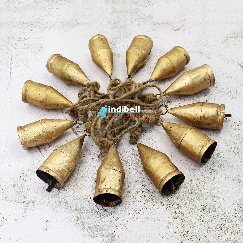 May include: A cluster of ten gold-colored metal bells, each with a conical shape, are strung together with a natural rope. The bells are arranged in a circular pattern, with the rope forming a central hub. The bells have a rustic, aged appearance.