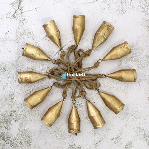 May include: A cluster of twelve gold-colored metal bells, each with a conical shape, are suspended from a central point by natural-colored rope. The bells are arranged in a circular pattern against a textured white background.