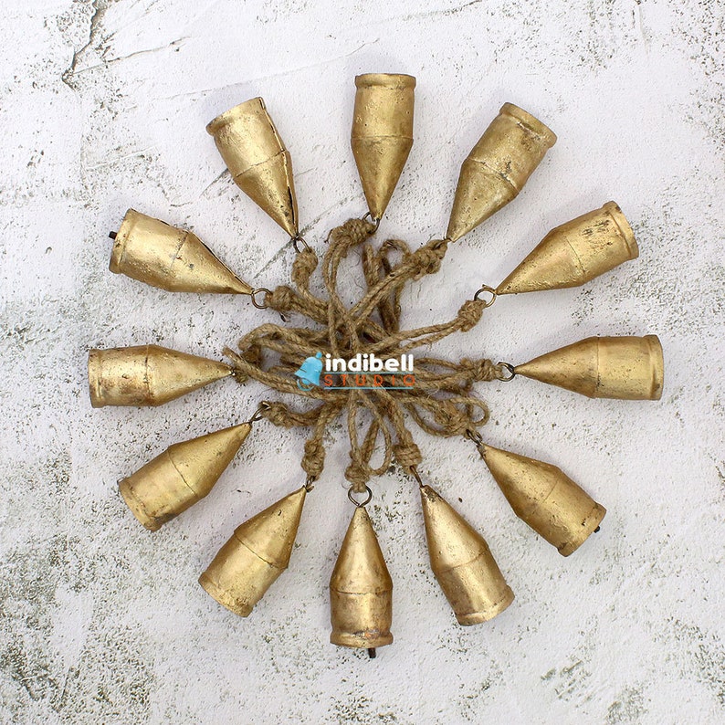 May include: A cluster of twelve gold-colored metal bells, each with a conical shape, are suspended from a central point by natural-colored rope. The bells are arranged in a circular pattern against a textured white background.