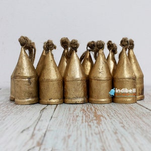 May include: A collection of golden-colored metal bells with a conical shape. Each bell has a loop of natural-colored rope at the top. The bells are arranged closely together on a light-colored wooden surface.