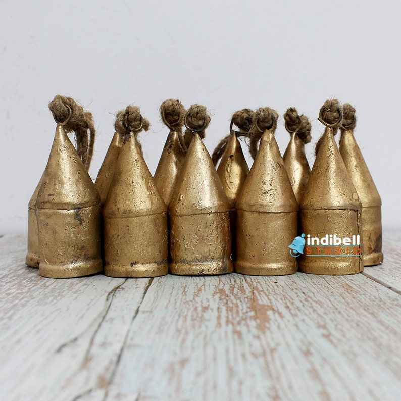 May include: A collection of golden-colored metal bells with a conical shape. Each bell has a loop of natural-colored rope at the top. The bells are arranged closely together on a light-colored wooden surface.