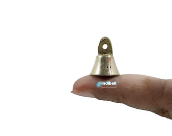 May include: A small, gold-coloured bell with a loop at the top. The bell is held on a finger against a white background. The bell has a conical shape and a small opening at the bottom.