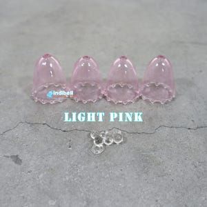 K&ouml;nnte beinhalten: Vier durchscheinende hellrosa glockenf&ouml;rmige Abdeckungen mit gewelltem Rand in einer Reihe angeordnet, begleitet von einer kleinen Ansammlung klarer kugelf&ouml;rmiger Perlen. Der Text indibell und LIGHT PINK ist sichtbar.