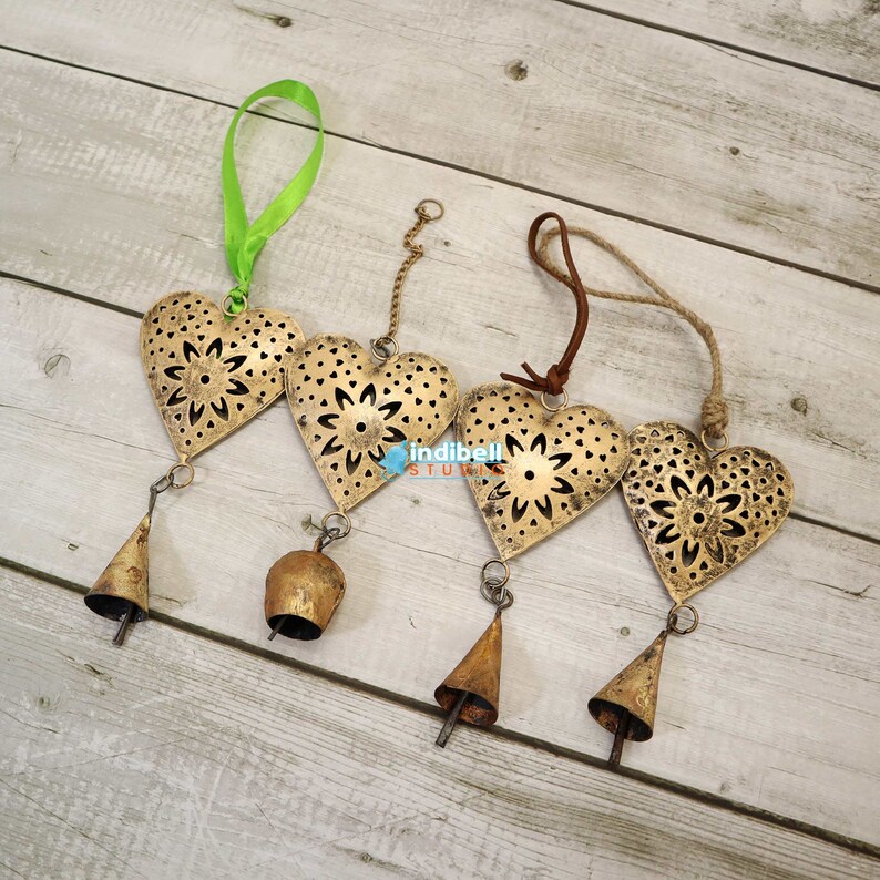 A set of four heart metal cutouts with vintage designs, each attached to bells and hung with various ribbons and strings.