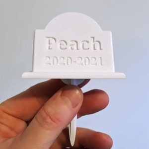 May include: White plastic pet memorial marker with the text "Peach 2020-2021" engraved on it. The marker has a pointed stake on the bottom for inserting into the ground.