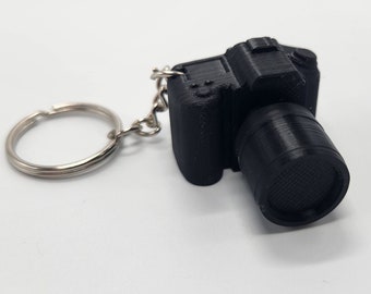May include: A black camera keyring with a silver ring and chain. The camera has a lens and a small button on top.
