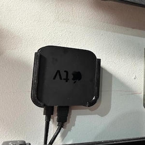 Puede incluir: Un dispositivo de streaming Apple TV negro montado en una pared blanca con dos cables negros conectados a la parte posterior. El Apple TV está etiquetado como "Apple TV" en texto blanco.