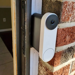 Può includere: Un campanello con telecamera bianco e nero montato su un muro di mattoni accanto a una porta.