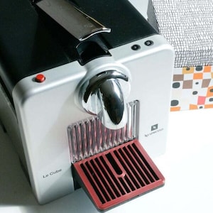 May include: A silver and black Nespresso coffee machine with a red drip tray. The machine has a chrome spout and a red button on the side. The text "Le Cube" is visible on the front of the machine.