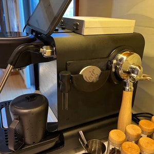 Op de afbeelding: Een zwarte espressomachine met een houten handvat en een zilveren portafilter. De machine heeft een digitaal display en een ingebouwde molen. Er staan ​​verschillende kleine potjes koffiebonen op het aanrecht onder de machine.