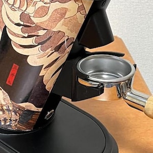 May include: A black and silver coffee grinder with a Japanese-inspired design featuring a skeletal figure and a wave pattern. The grinder has a portafilter attached to the spout.
