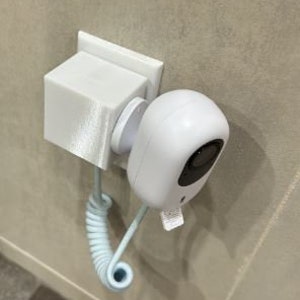 May include: A white wireless security camera with a coiled cord attached to a white wall mount. The camera is mounted on a wall outlet.