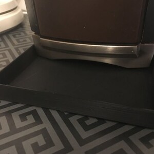 Puede incluir: Un objeto decorativo marrón oscuro y negro con una banda plateada, apoyado sobre una bandeja cuadrada negra. La bandeja tiene una superficie ligeramente texturizada. El objeto es probablemente un artículo decorativo.