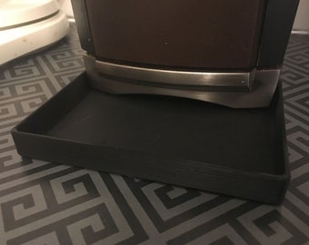 Puede incluir: Un objeto decorativo marrón oscuro y negro con una banda plateada, apoyado sobre una bandeja cuadrada negra. La bandeja tiene una superficie ligeramente texturizada. El objeto es probablemente un artículo decorativo.