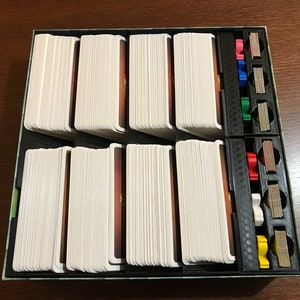 May include: A game organizer with multiple compartments for storing cards and tokens. The organizer is made of black plastic and has a green and brown patterned design. The organizer is filled with white cards and colored tokens.