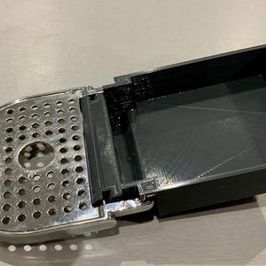 Puede incluir: Un componente metálico plateado perforado está unido a un contenedor rectangular negro. La pieza metálica tiene una forma redondeada con numerosos agujeros pequeños. El contenedor está abierto, revelando su interior.