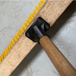 May include: A close-up of a wooden broom handle with a black plastic connector and yellow bristles. The broom handle is made of wood and has a natural finish. The connector is made of black plastic and has four screws.