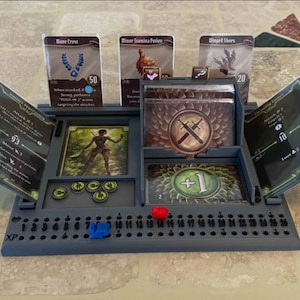 Puede incluir: Un tablero de juego de plástico gris con una variedad de cartas y fichas. El tablero tiene una cuadrícula de números y una sección para cartas. También hay varias fichas, incluyendo una roja, una azul y varias verdes.