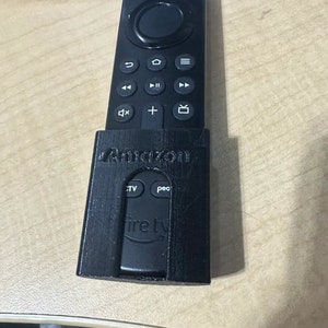 Puede incluir: Un mando a distancia negro con una funda negra. El mando tiene botones para encender, inicio, menú, volumen, reproducir, pausar, avanzar rápido, retroceder y un botón con un icono de TV. La funda tiene la palabra "Amazon" impresa.