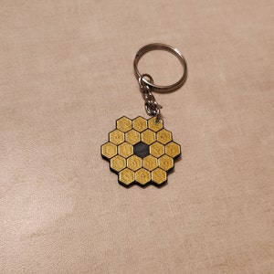 May include: A gold-coloured, hexagonal keychain charm with a honeycomb design, attached to a silver chain and ring. The centre of the charm is a dark grey colour. The keychain is a space-themed accessory.