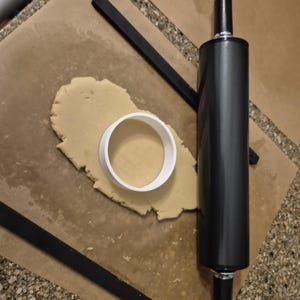 May include: A black rolling pin and a white cookie cutter on a brown surface with a sheet of parchment paper. The cookie cutter is round and the rolling pin is about 12 inches long.