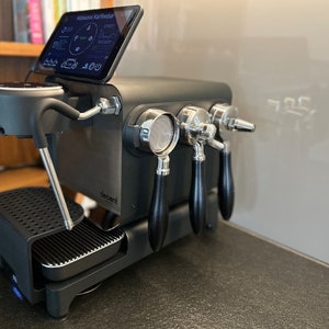 Puede incluir: Una máquina de espresso negra y plateada con una pantalla táctil que dice "Willkommen Kaffeebar". La máquina tiene dos boquillas y un portafiltro.