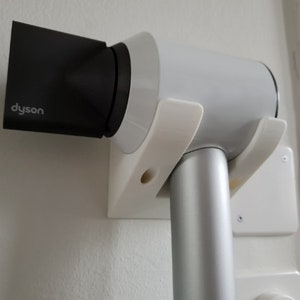 May include: White wall-mounted holder with a silver Dyson hairdryer attached. The holder has two prongs that hold the hairdryer in place. The hairdryer has a black nozzle.