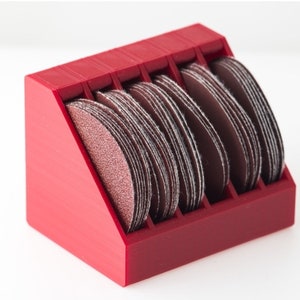 May include: Red plastic storage organizer with multiple compartments for holding sanding discs. The organizer is filled with brown sanding discs.