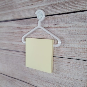 May include: A white plastic hanger with a yellow sticky note attached to it. The hanger is mounted on a wooden wall.