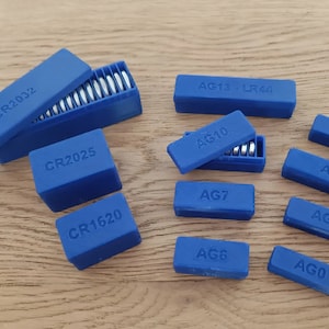 Puede incluir: Surtido de contenedores de almacenamiento de baterías de plástico azul etiquetados con diferentes tipos de baterías, incluyendo CR2032, CR2025, CR1620, AG13 LR44, AG10, AG7, AG6, AG4, AG3, AG1 y AG0.
