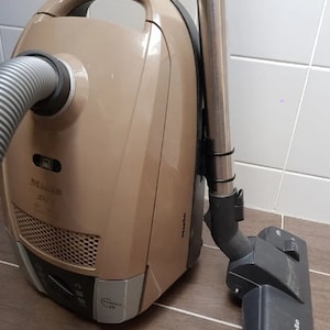 以下が含まれることがあります： ベージュの縦型掃除機に、シルバーの延長ワンドとブラックの床用ノズルが付いています。掃除機はブラックのハンドルとシルバーの電源スイッチが付いています。床用ノズルには「Miele」のブランド名が印刷されています。