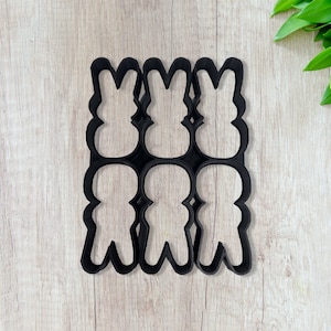 May include: Black plastic cookie cutter set with six bunny shapes. The cutter has a rectangular frame with three bunny shapes on top and three on the bottom. The bunnies have long ears and rounded bodies. The cutter is on a light wood surface.
