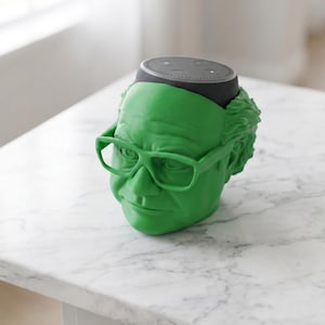 May include: A green decorative speaker holder shaped like a head with glasses. The top of the speaker is black and sits in the head. The holder is on a white marble table.