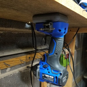May include: A blue and black cordless drill with a battery attached is hanging from a wooden shelf. The drill is hanging upside down with the drill bit facing down. The drill is attached to the shelf with a magnetic holder.