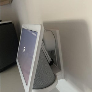 Peut inclure: Un écran intelligent Google Nest Hub Max blanc monté sur un mur avec une plaque de prise blanche. L'écran affiche le logo Google. L'appareil est connecté à une prise de courant avec plusieurs cordons.