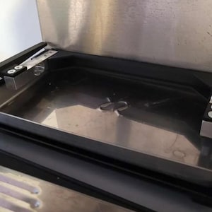 May include: Stainless steel appliance interior with a rectangular tray and a curved indentation. The tray is surrounded by a black frame and secured with metal brackets and screws. The appliance has a metallic finish.